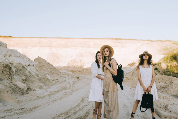 beautiful stylish girlfriends in elegant dresses and straw hats walking in sandy canyon - Photo, Image