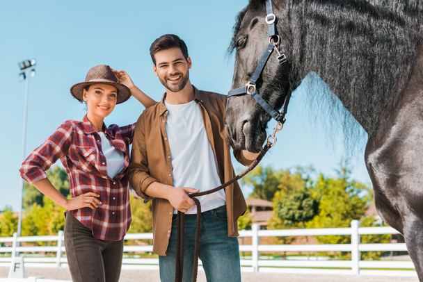 hembras sonrientes y equestrians masculinos de pie cerca del caballo en el rancho y mirando a la cámara
 - Foto, imagen