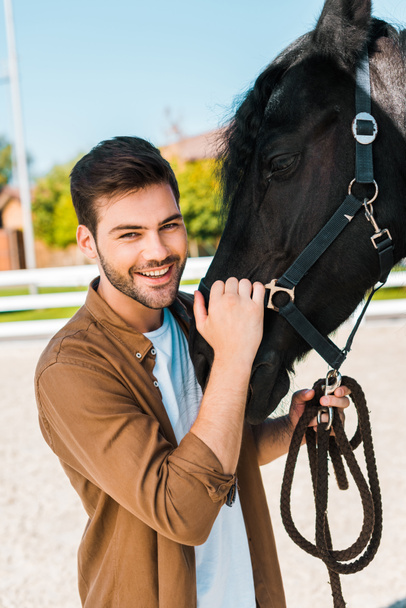 smiling handsome male equestrian holding horse halter and looking at camera at ranch - Photo, Image