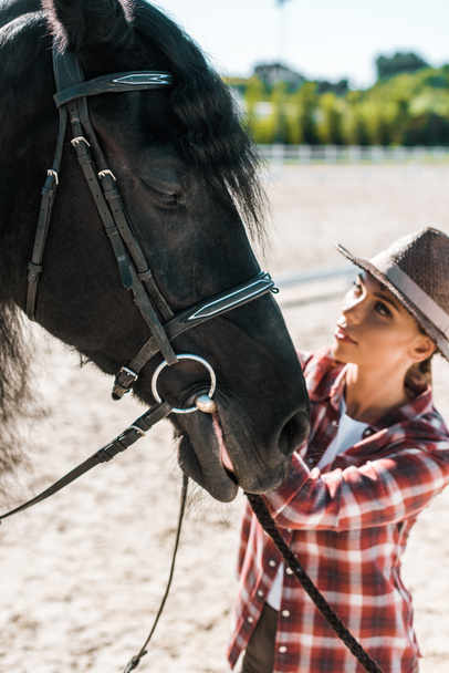 beautiful female equestrian in checkered shirt and hat fixing horse halter at ranch - Photo, Image