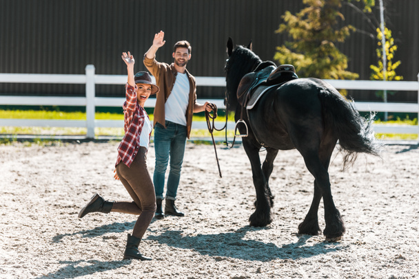 счастливые наездники женского и мужского пола, идущие с лошадью на ранчо и машущие руками
 - Фото, изображение