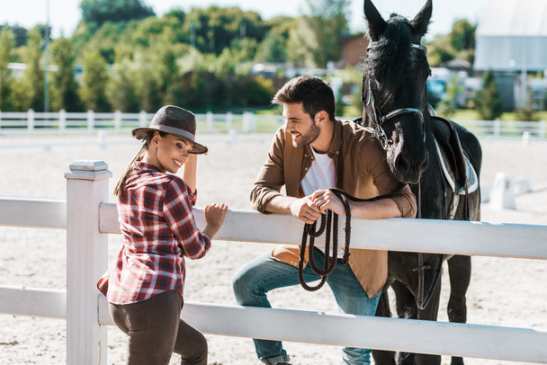Lächelnder Cowboy und Cowgirl stehen am Zaun mit Pferd und unterhalten sich auf Ranch - Foto, Bild