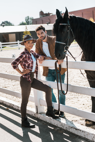 cavaliers féminins et masculins debout près de la clôture avec cheval au ranch et regardant la caméra
 - Photo, image