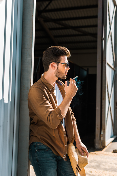 side view of handsome man in sunglasses smoking cigarette and leaning on wall at ranch - Photo, Image
