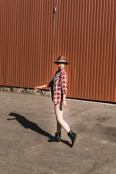 attractive cowgirl in checkered shirt and hat walking near brown building at ranch - Photo, Image