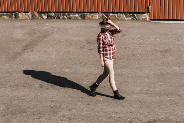 attractive woman in checkered shirt touching hat and walking near brown building at ranch - 写真・画像