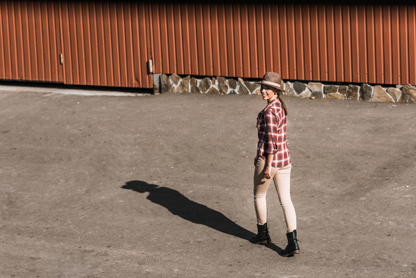 smiling attractive cowgirl in checkered shirt and hat walking near brown building at ranch - Zdjęcie, obraz