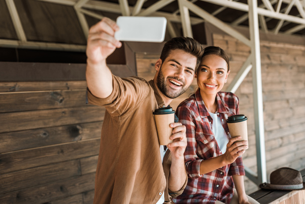 sorrindo homem e mulher tomando selfie com smartphone e segurando copos de café descartáveis no rancho
 - Foto, Imagem