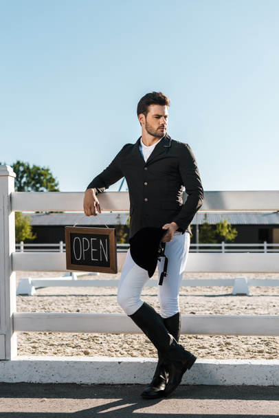 handsome male equestrian leaning on fence and holding open sign at horse club - Photo, Image