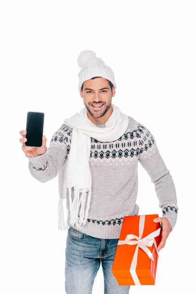 schöner junger Mann in Pullover, Schal und Mütze mit Geschenkbox und Smartphone mit leerem Bildschirm auf weißem Hintergrund - Foto, Bild
