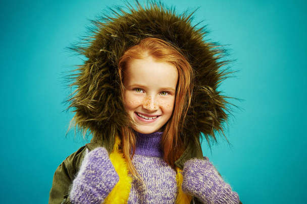 Portrait of beautiful children girl cute smiling, wears autumn warm jacket with fur hood, expresses sincerity, has red hair and freckles. Prepared for cold child. - Photo, Image