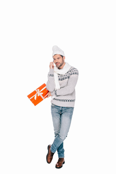 smiling young man in sweater, scarf and hat holding gift box and talking by smartphone isolated on white  - Zdjęcie, obraz