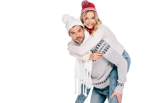 beautiful happy young couple in sweaters, scarves and hats piggybacking and smiling at camera isolated on white - Fotografie, Obrázek