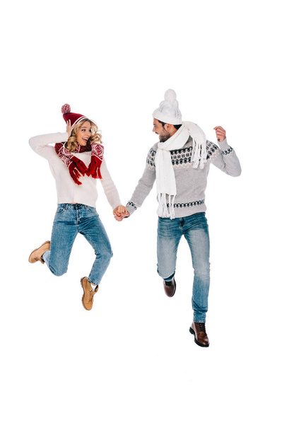 feliz joven pareja en suéteres y sombreros tomados de la mano y saltando aislado en blanco
 - Foto, Imagen