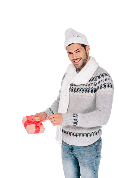 handsome young man in sweater, scarf and hat opening gift box and smiling at camera isolated on white - Фото, изображение