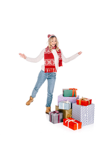 όμορφη happy νεαρή γυναίκα σε κασκόλ και καπέλο βλέπουν κιβώτια στοιβάζονται δώρων που απομονώνονται σε λευκό - Φωτογραφία, εικόνα