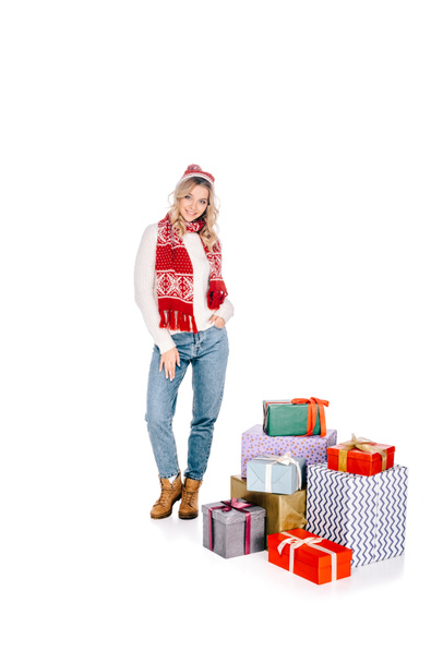 beautiful young woman in scarf and hat standing near stacked gift boxes and smiling at camera isolated on white  - Zdjęcie, obraz