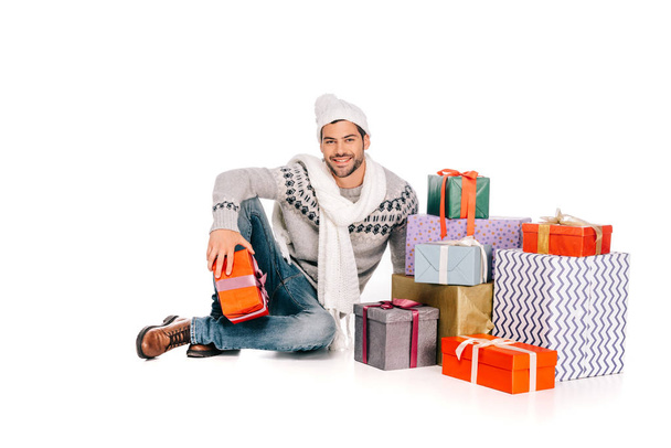 hombre guapo en bufanda y sombrero sentado cerca de cajas de regalo y sonriendo a la cámara aislado en blanco
 - Foto, Imagen