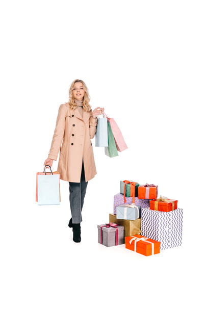 beautiful young woman holding shopping bags and looking at camera while standing near gift boxes isolated on white - Photo, Image