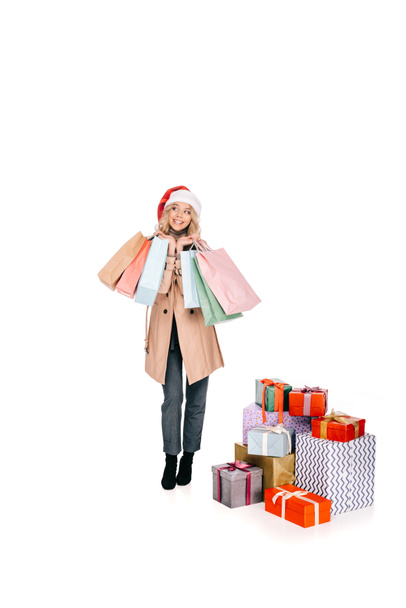 schöne lächelnde junge Frau mit Weihnachtsmannmütze, Einkaufstüten in der Hand und in der Nähe von Geschenkboxen isoliert auf weiß - Foto, Bild
