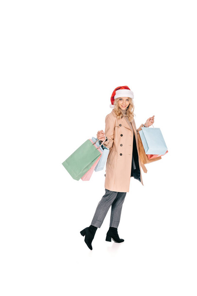 full length view of beautiful happy young woman in santa hat holding shopping bags and smiling at camera isolated on white  - Foto, Imagen