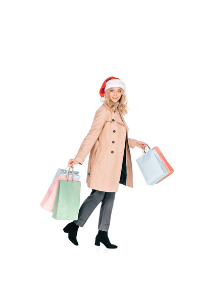 full length view of woman in santa hat walking with shopping bags and smiling at camera isolated on white - Foto, immagini