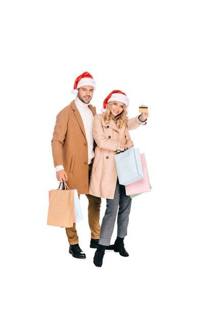gelukkige jonge paar in santa hoeden holding creditcard en boodschappentassen geïsoleerd op wit  - Foto, afbeelding