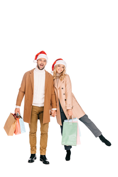 beautiful happy young couple in santa hats holding shopping bags and smiling at camera isolated on white  - Foto, Bild
