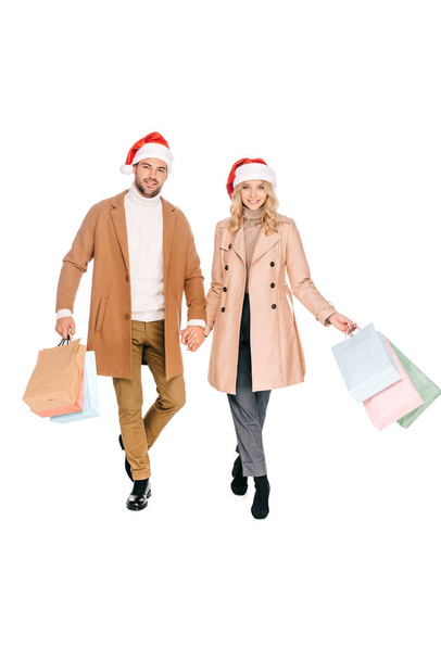 happy young couple in santa hats holding shopping bags and smiling at camera isolated on white  - Фото, зображення