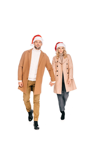 Elele ve üzerinde beyaz izole kamera gülümseyen Noel Baba şapkaları güzel genç çift - Fotoğraf, Görsel
