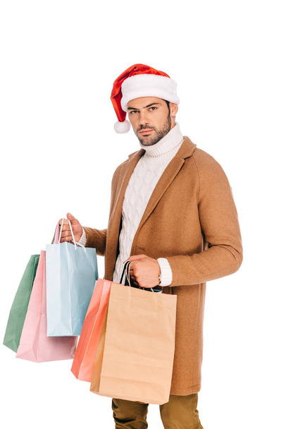 handsome young man in santa hat holding shopping bags and looking at camera isolated on white - Fotó, kép