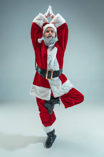 Funny serious guy with christmas hat sitting at yoga pose at studio. New Year Holiday. Christmas, x-mas, winter, gifts concept. Man wearing Santa Claus costume on gray. Copy space. Winter sales. - Fotó, kép