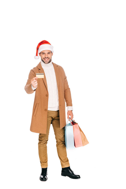 usměvavý mladý muž v santa hat držení platební karty a nákupní tašky izolované na bílém - Fotografie, Obrázek