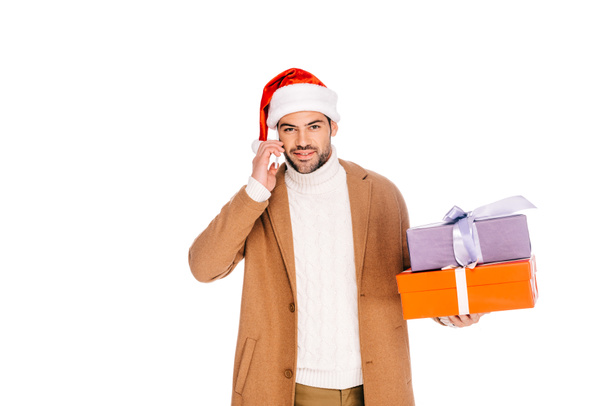 joven en santa hat sosteniendo cajas de regalo y hablando por teléfono inteligente aislado en blanco
 - Foto, imagen