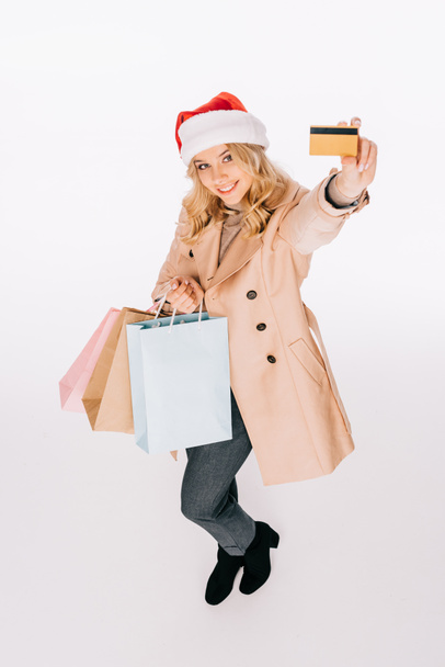 высокий угол зрения счастливой молодой женщины в шляпе Санта держа сумки и кредитные карты изолированы на белом
 - Фото, изображение