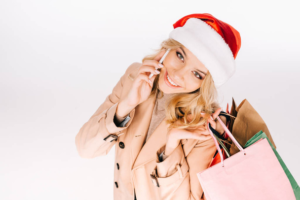pohled z vysokého úhlu krásné usměvavé mladé ženy v santa hat drží nákupní tašky a mluví o smartphone izolované na bílém - Fotografie, Obrázek