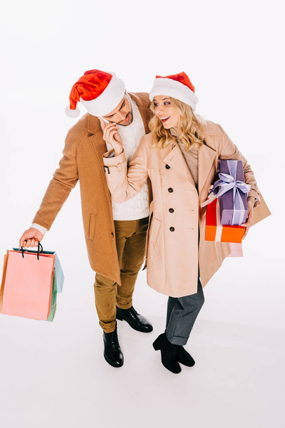 μεγάλη γωνία προβολής του happy νεαρό ζευγάρι σε καπέλα santa κρατώντας δώρο κουτιά και τσάντες αγορών που απομονώνονται σε λευκό - Φωτογραφία, εικόνα