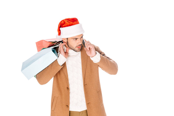 νεαρός άνδρας με το καπέλο santa, κρατώντας τσάντες για ψώνια και να μιλάμε από smartphone που απομονώνονται σε λευκό - Φωτογραφία, εικόνα