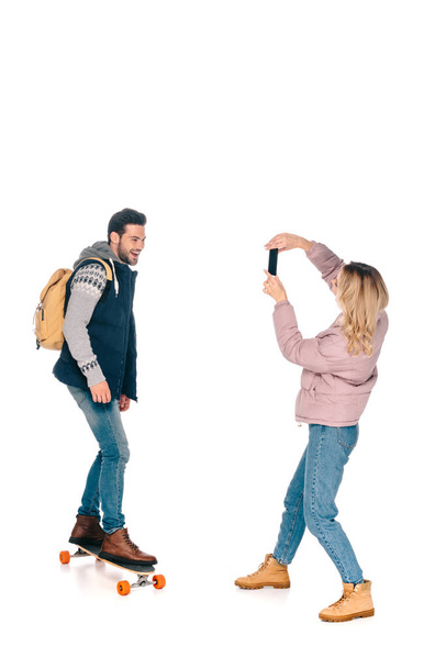 mujer joven con smartphone fotografiando hombre sonriente con mochila montando longboard
  - Foto, Imagen