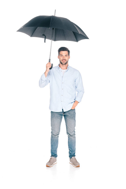 full length view of handsome young man holding black umbrella and looking at camera isolated on white - Foto, Imagem