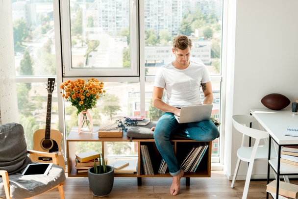 Nuori mies rento vaatteet kannettavan tietokoneen kun istuu ikkunassa kotona
 - Valokuva, kuva