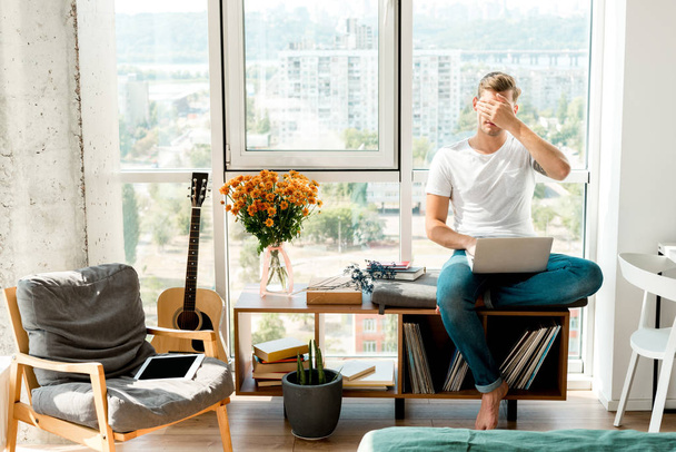 alkalmi ruházat, laptop, amely szemét, miközben ül otthon ablakban fiatalember - Fotó, kép