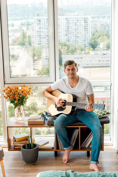 young guitarist playing acoustic guitar while sitting at window at home - Фото, зображення