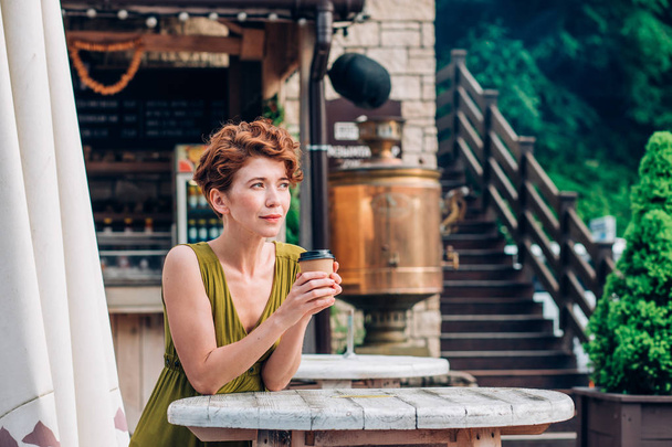 A beautiful redhead woman in a green dress stands, holds coffee in her hands and looks away with a smile. Summer, relax, vacation concept - Zdjęcie, obraz