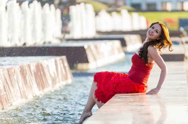 Beautiful young girl in red walks through the spring city walking by the fountain - Foto, Imagen