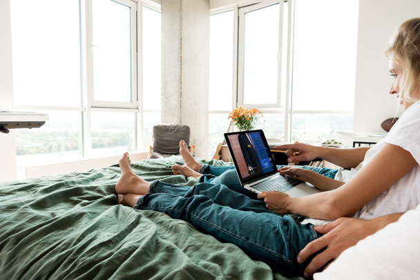 vista parcial de la pareja usando portátil digital con signo de reserva en la pantalla mientras descansa en la cama en casa
 - Foto, imagen