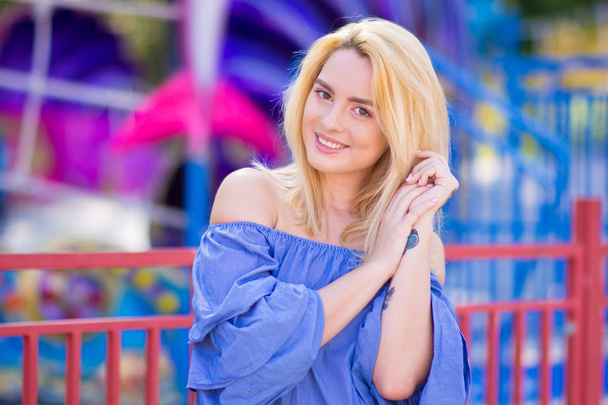 Beautiful young woman with blond hair smiling on a bright background of blurred amusement park - Zdjęcie, obraz