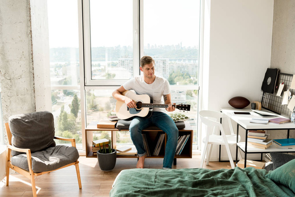 jovem em roupas casuais tocando guitarra acústica em casa
 - Foto, Imagem