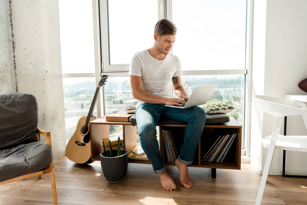 young man in casual clothing using laptop while sitting at window at home - Foto, imagen