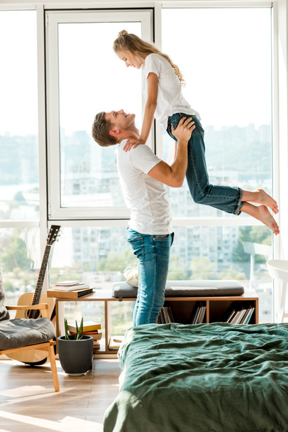 side view of cheerful man holding girlfriend in hands while standing at window at home - Fotoğraf, Görsel
